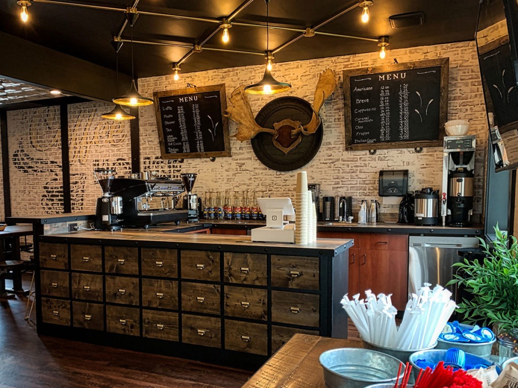 A bistro with brown wooden kitchen cabinets with kitchen tools over wooden counters. The floor is also of wood. There is a rich brown ceiling with task and ambient lighting.